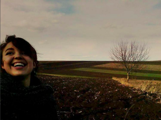 Young woman's face on the left side, freshly tilled field immediately behind her and fields just turning green beyond that, single bare tree in the midground on the right.